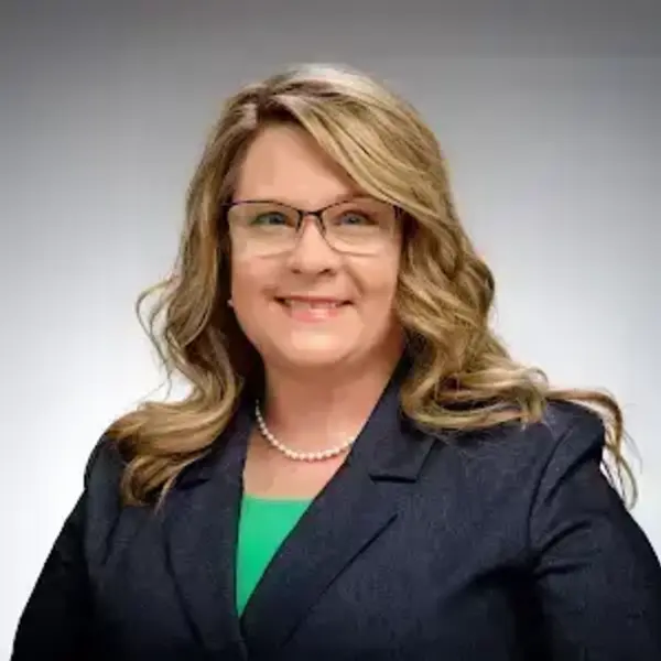 Woman with blonde wavy hair, glasses, and a bright smile. She wears a navy blazer, green top, and pearl necklace.